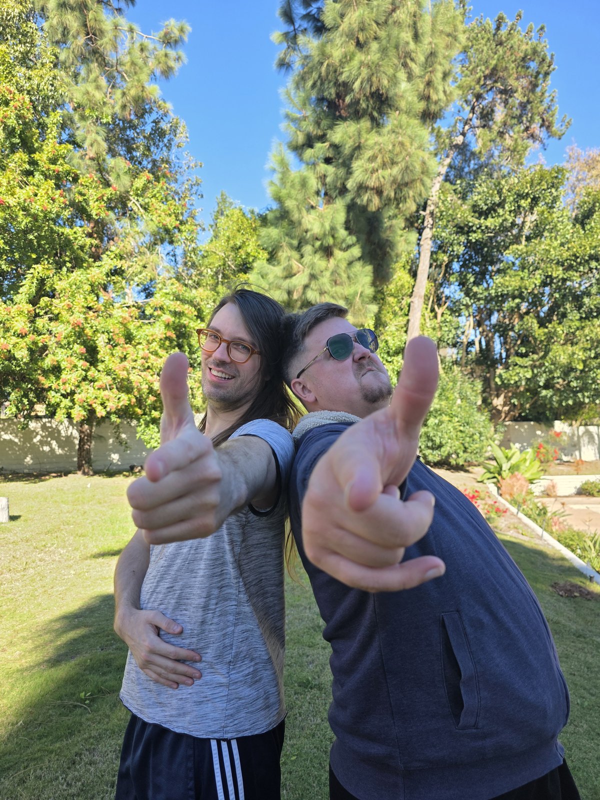Goose and Squirrelybass back-to-back pointing finger guns at the camera in a sunny backyard