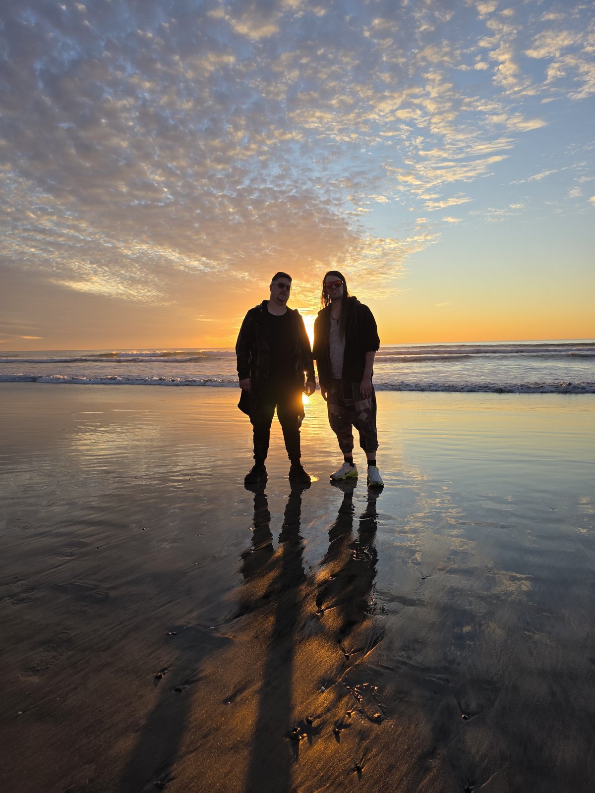 Goose and Squirrelybass standing in shallow surf at sunset, the sun between them, reflected in the wet sand